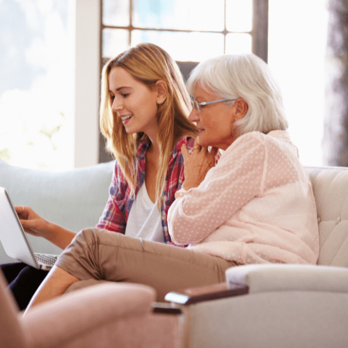 Learning abouta proprietary reverse mortgage An older woman with white shoulder length hair wearing a light pink cardigan and khaki pants and a younger woman with blond mid length hair wearing a plaid shirt open over a white cami tank top sitting together on a beige sofa with a laptop. They are both looking at the laptop intently.