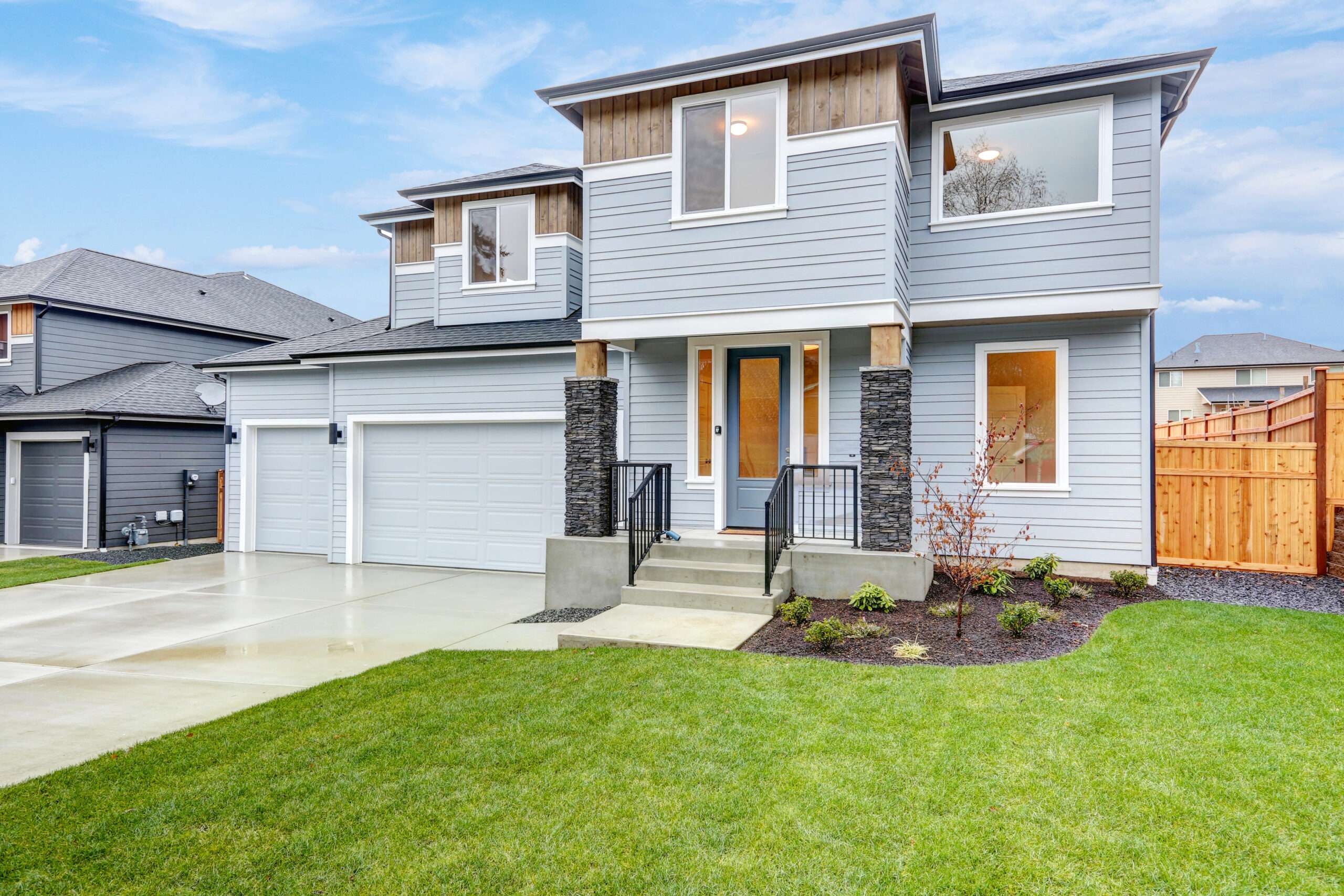 Modern two-story house with gray siding, large windows, and a double garage. The well-manicured lawn and clear blue sky create a welcoming, serene atmosphere.